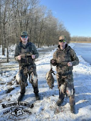 Two people in camouflage gear stand on snow by water, each holding several ducks they have hunted. Leafless trees are in the background; a "Waterfowl Unlimited" logo appears in the corner.