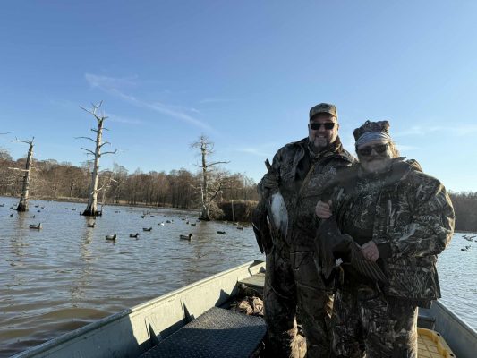 Two people in camouflage stand on a boat holding ducks, surrounded by water, decoy ducks, and leafless trees under a clear sky.