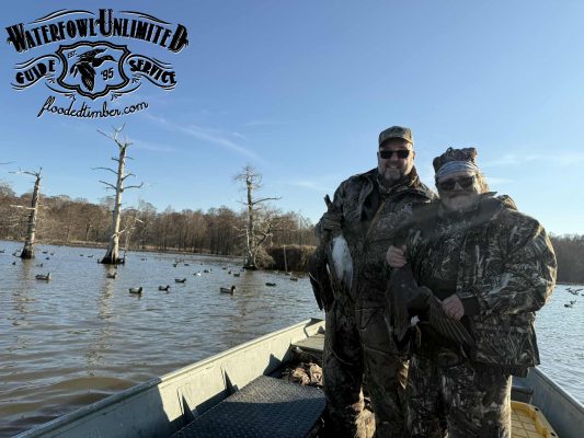 Two people in camouflage pose on a boat with ducks in hand on a flooded timber lake, surrounded by decoys and leafless trees. "Waterfowl Unlimited Guide Service" logo is in the corner.