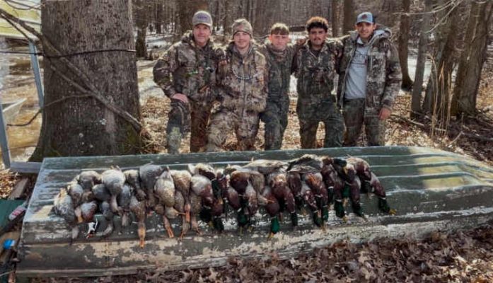 Five people in camouflage clothing stand behind a row of harvested ducks laid out on an overturned boat in a wooded area.