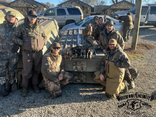 Seven people in camouflage and waders pose around a metal table with hunted ducks displayed on top; cabins and vehicles are in the background.
