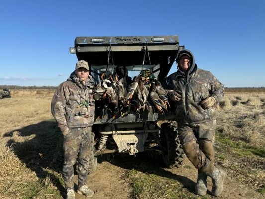 Two people in camouflage clothing stand next to an ATV with several ducks hanging from the back, in a grassy field under a clear sky.
