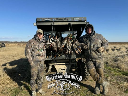 Two people in camouflage stand by an ATV loaded with hunted ducks in a field. The ATV displays a "Waterfowl Unlimited Guide Service" logo on the tailgate.