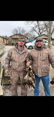 Two men in camouflage jackets stand outdoors; one holds several ducks by their feet. A boat, cars, and houses are visible in the background.