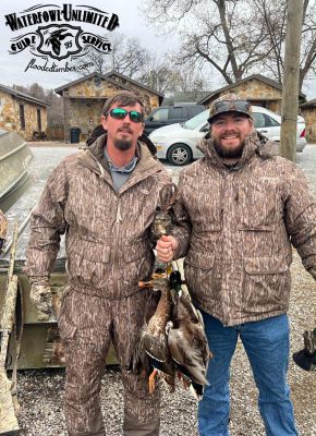 Two men in camouflage jackets stand outdoors holding several ducks, with cabins and a truck in the background. A “Waterfowl Unlimited Guide Service” logo appears in the corner.