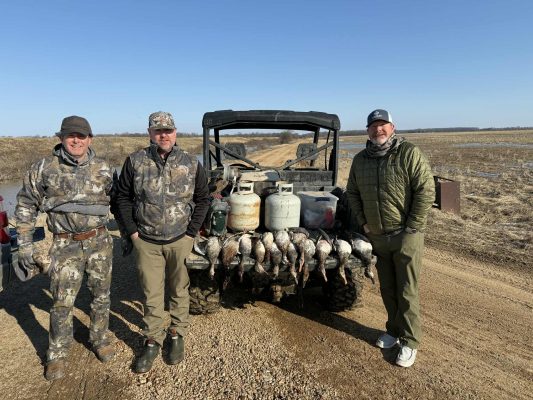 Three men in outdoor clothing stand by an off-road vehicle with several propane tanks and a row of harvested ducks displayed on the back, on a dirt road near a marshy field.