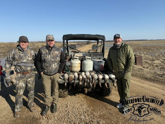 Three men in hunting gear stand next to an ATV with waterfowl and propane tanks on the back in an open field. "Waterfowl Unlimited" logo is visible in the lower right corner.