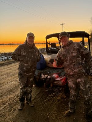 Two people in camouflage clothing stand by an off-road vehicle at sunset, with game animals loaded in the back and gear bags nearby on a dirt road.