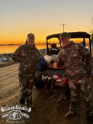 Two people in camouflage gear stand next to an ATV loaded with gear and game birds at sunset on a dirt road, with the "Waterfowl Unlimited Guide Service" logo in the corner.