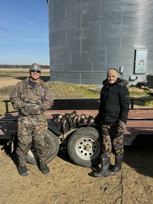 Two people in outdoor gear stand beside a trailer displaying several ducks, with a large metal grain bin in the background.