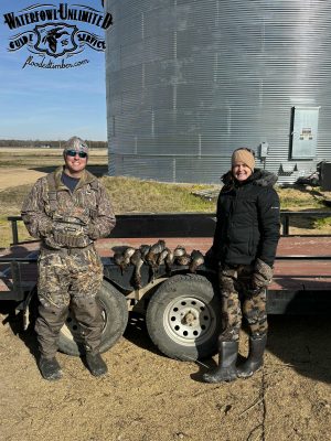Two people in camouflage clothing stand in front of a trailer displaying hunted ducks, with a large metal grain silo in the background.