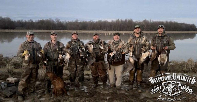 Six hunters in camouflage stand by a lakeshore holding ducks, with a dog sitting at their feet; Waterfowl Unlimited Guide Service logo is in the bottom right corner.