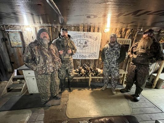 Four men in camouflage hunting gear stand indoors in front of a "Waterfowl Unlimited" sign, with a display of ducks on the floor below the sign.
