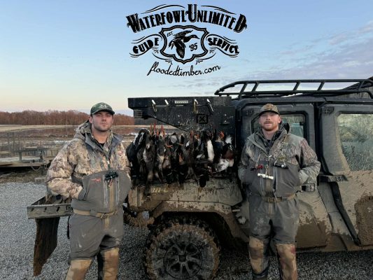 Two men in camo hunting gear stand beside a muddy off-road vehicle with harvested ducks hanging on it; "Waterfowl Unlimited Guide Service" logo is displayed above.