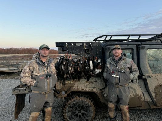 Two men in camouflage gear stand beside a muddy off-road vehicle with a row of ducks hanging from the back, in an outdoor setting at sunrise or sunset.