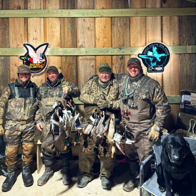 Four hunters in camouflage pose indoors with harvested ducks; a black dog sits on a chair nearby. Logos for Ducks Unlimited and Black Duck Revival are on the wooden wall behind them.