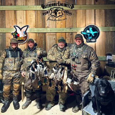 Four hunters in camouflage pose indoors with hunted ducks hanging from straps. A black dog sits nearby. Hunting club signs decorate the wooden wall behind them.