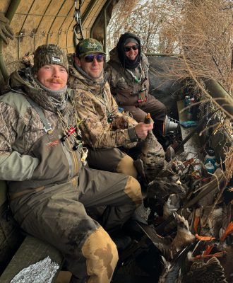 Three people in camouflage gear sit in a hunting blind, holding ducks with more ducks at their feet. They are surrounded by dry branches and cold weather gear.