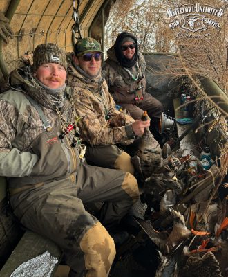 Three men in camouflage hunting gear sit inside a hunting blind with several ducks and geese laid out in front of them. "Waterfowl Unlimited Guide Service" logo is visible in the corner.