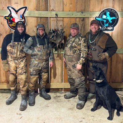 Four men in camouflage hunting gear stand indoors by a wooden wall with hunting signs, posing with a black dog and several hunted birds hanging behind them.