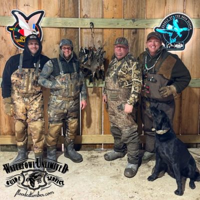 Four men in camouflage gear stand indoors with a black dog and a group of hanging ducks; wooden wall and hunting club logos are visible in the background.