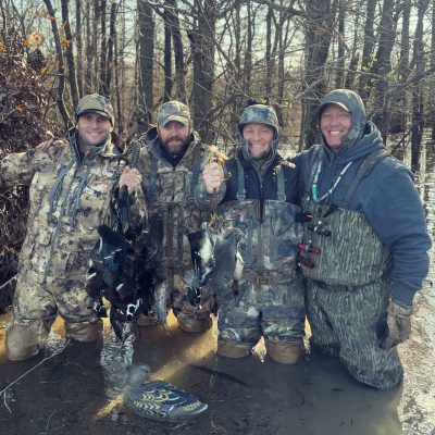 Four men in camouflage waders stand in shallow water in a wooded area, holding several ducks they have hunted; a duck decoy floats in front of them.