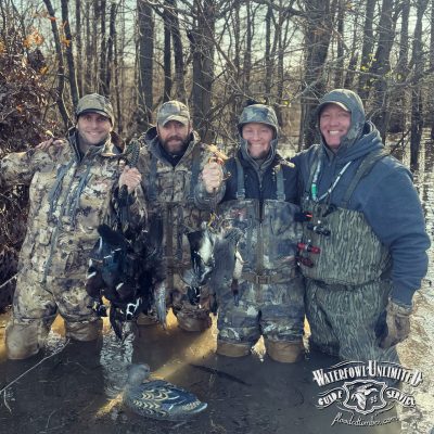 Four people in camouflage and waders stand in shallow water in a wooded area, holding up several ducks. A decoy and "Waterfowl Unlimited Guide Service" logo are visible in the foreground.