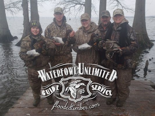 Five people in camouflage clothing stand on a dock holding ducks, with trees and water in the background. "Waterfowl Unlimited Guide Service" logo is overlaid on the image.