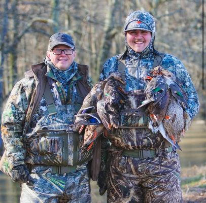 Two people in camouflage gear stand outdoors by water, smiling and holding several ducks from a hunt.