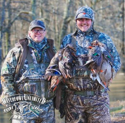 Two hunters in camouflage clothing stand outdoors, one holding several ducks. A "Waterfowl Unlimited Guide Service" logo is visible in the lower left corner.