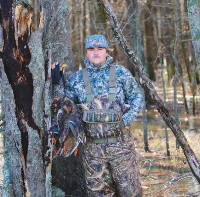 A man in camouflage hunting gear stands in a forest holding ducks by his side, with trees and water in the background.