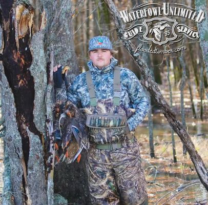 A man in camouflage hunting gear stands in a flooded forest holding several ducks. The "Waterfowl Unlimited Guide Service" logo is displayed in the top right corner.