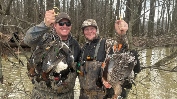 Two hunters in camouflage and waders stand in shallow water, each holding several ducks they have hunted, surrounded by leafless trees.