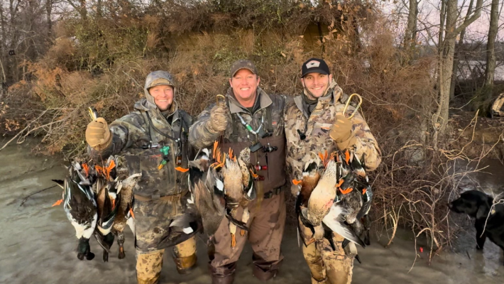 Three people in camouflage clothing stand outdoors, smiling and holding several ducks they have hunted, with brush and trees in the background.