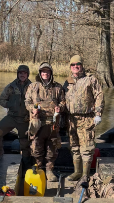 Three people in camouflage clothing stand on a boat holding ducks, with leafless trees and a riverbank in the background. It is a bright, sunny day.