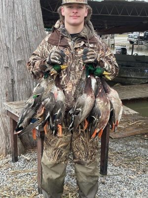 A person in camouflage clothing stands outdoors holding several hunted ducks by their necks, with boats and trees in the background.