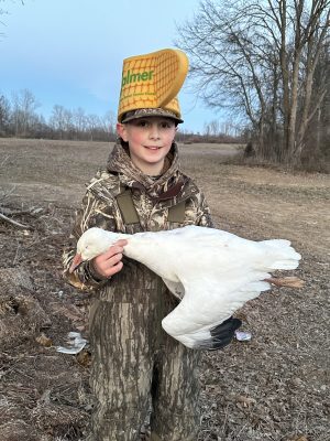 A child in camouflage clothing holds a large white bird and wears a foam hat shaped like a cheese wedge with “Palmer” printed on it, standing outdoors in a field near leafless trees.