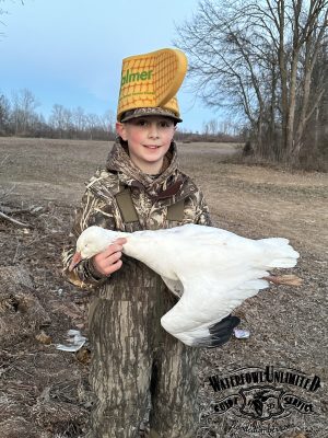 A child in camouflage clothing and a novelty foam hat holds a white goose. The background shows a bare field with some trees and scattered debris.