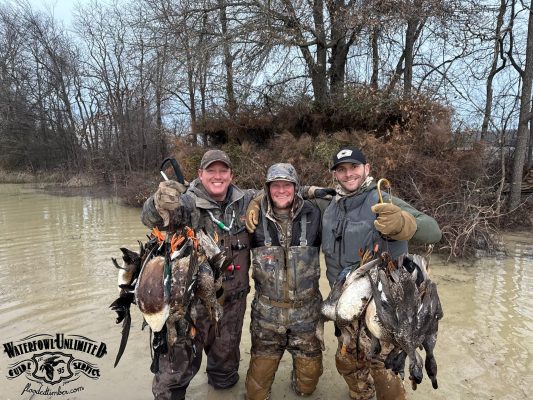 Three people in hunting gear stand in shallow water, smiling and holding multiple ducks. Leafless trees and muddy terrain are visible in the background. Waterfowl Unlimited logo is present.