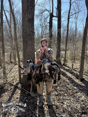 A person in camouflage hunting gear stands in a forest, holding several harvested ducks. Sunlight filters through leafless trees. The "Waterfowl Unlimited Guide Service" logo appears in the lower left.