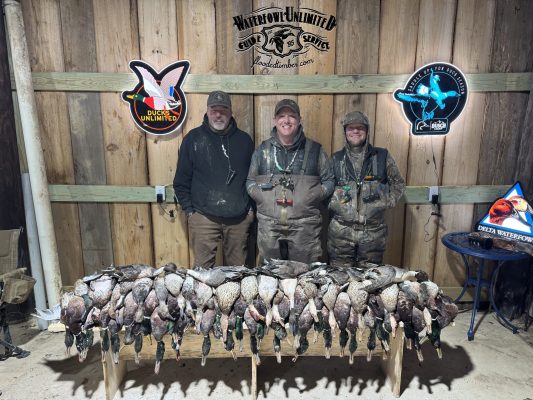 Three people in hunting gear stand behind a table displaying numerous ducks in a wooden room, with hunting-themed signs on the wall behind them.