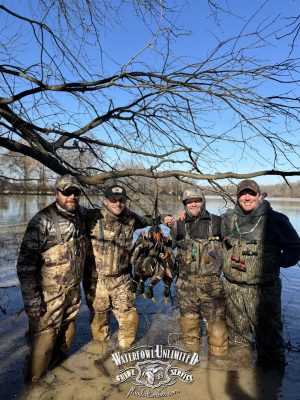 Four people in camouflage waders stand in shallow water, posing with a bundle of ducks under a leafless tree. The Waterfowl Unlimited logo is visible at the bottom of the image.