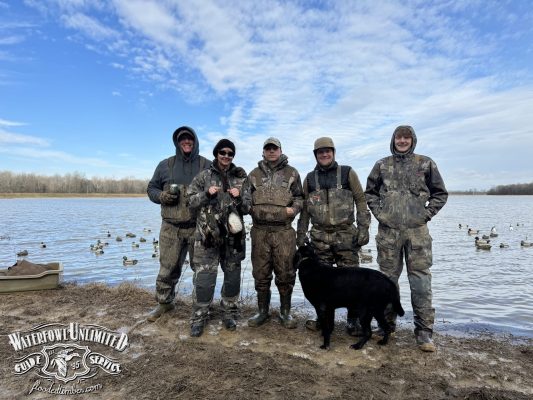 Five people in camouflage hunting gear stand by a lakeshore with decoys, holding ducks, and a black dog is in front of them. Waterfowl Unlimited Guide Service logo appears in the corner.