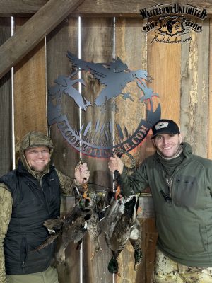 Two men in outdoor gear smile and hold several ducks by their feet in front of a wooden wall with a "Flooded Timber Waterfowl Unlimited Guide Service" logo.
