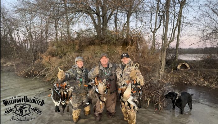 Three hunters in camouflage stand on icy ground holding several ducks, with trees, brush, and a black dog nearby. A Waterfowl Unlimited Guide Service logo appears in the lower left corner.