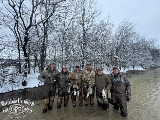 Six people in camouflage gear stand in shallow, snowy water holding ducks, with bare trees and snow-covered ground in the background. "Whitefowl Unlimited Guide Service" logo in bottom left corner.