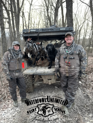 Two hunters in camouflage stand beside an ATV with a dog and several harvested ducks in a wooded area. "Waterfowl Unlimited Guide Service" logo is visible at the bottom.