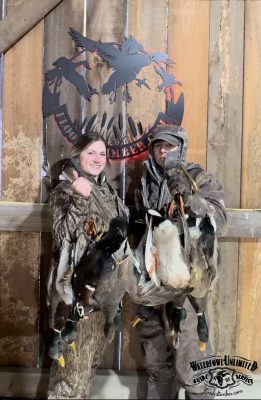 Two people in camouflage hold several ducks inside a wooden structure, with a "Waterfowl Unlimited" sign featuring duck graphics in the background.