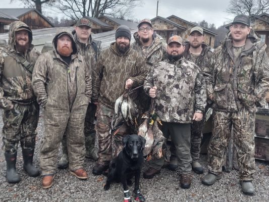 Nine men in camouflage hunting gear stand outdoors with a black dog; two men hold several ducks, and cabins are visible in the background.