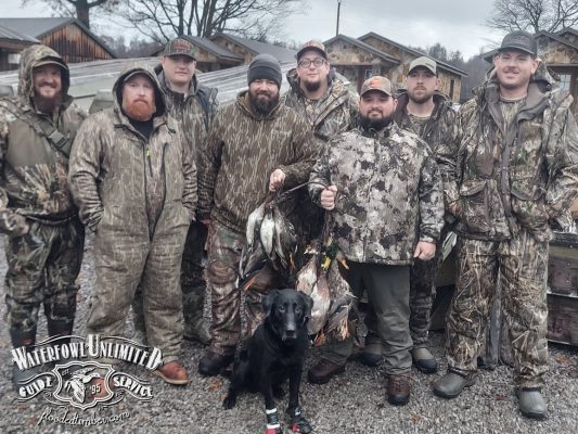 A group of eight men in camouflage clothing pose outdoors with a black dog and a collection of hunted birds; cabins and trees are in the background.